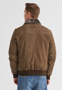 Braune Wildleder-Bomberjacke mit einem Kragen aus Kunstpelz, gerippten Bündchen und Saum sowie einem quadratischen Aufnäher auf dem Rücken. Jeanshose am unteren Rand sichtbar.