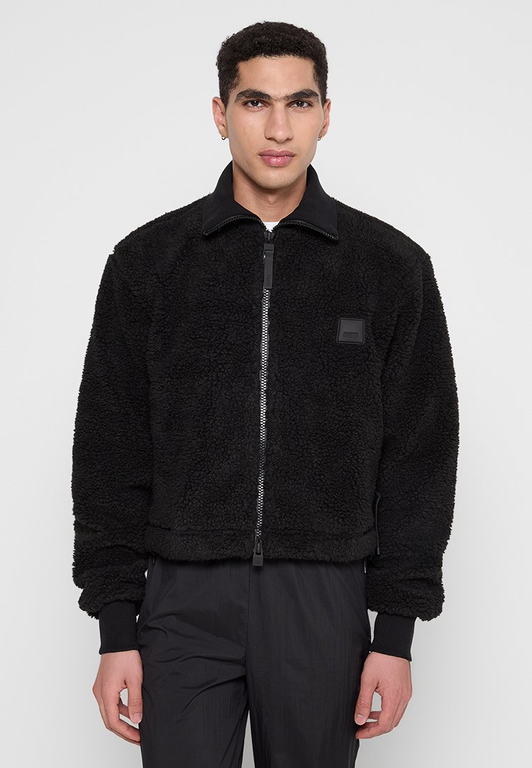 Veste en polaire noire avec un col haut, fermeture éclair à l'avant et poignets côtelés. Présente une surface texturée et un petit écusson logo.
