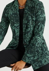 Grön mönstrad kavaj med bladmönster, med en lätt texturerad tyg, snibbade slag och en enkel knappstängning. Svart topp under.