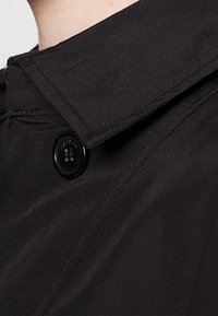 Trench-coat en tissu noir avec un col pointu, doté d'une fermeture à boutons et de détails de surpiqûres visibles pour une texture supplémentaire.