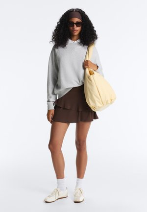 Nő napszemüvegben, szürke pulóverben, barna réteges szoknyában, fehér tornacipőben és zokniban, vállán egy nagy, halványsárga vászontáskát tartva.