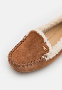 Brune semsket tøffel med kremfarget pelskant, dekorativ stikning og en gulllogo på siden. Flat, rund tådesign.