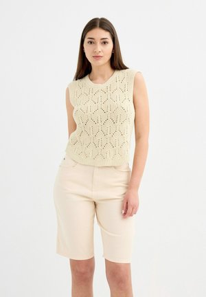 Mujer de pie, vistiendo una blusa sin mangas de punto beige con un patrón de encaje y pantalones cortos beige hasta la rodilla, frente a un fondo blanco.