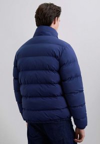 Hombre vestido con una chaqueta acolchada azul marino de cuello alto, de pie de espaldas contra un fondo claro y uniforme.