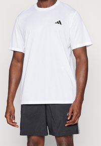 Camiseta deportiva blanca hecha de tejido ligero, con cuello redondo, mangas cortas y un logo negro en el lado izquierdo del pecho.