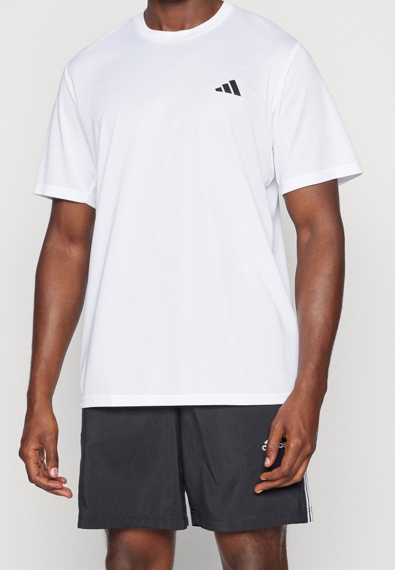Camiseta deportiva blanca hecha de tejido ligero, con cuello redondo, mangas cortas y un logo negro en el lado izquierdo del pecho.