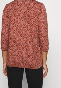 Blusa color ruggine con motivo paisley e floreale, maniche lunghe arrotolate fino ai gomiti e vestibilità ampia, realizzata in tessuto leggero.