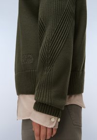 Manica di un maglione verde lavorato a maglia con un motivo a coste sopra il polsino di una camicia beige e pantaloni grigi, visibili su un braccio e una mano.