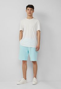 Weißes Baumwoll-T-Shirt mit kurzen Ärmeln, kombiniert mit hellblauen Shorts und weißen Sneakern, steht vor einem schlichten grauen Hintergrund.