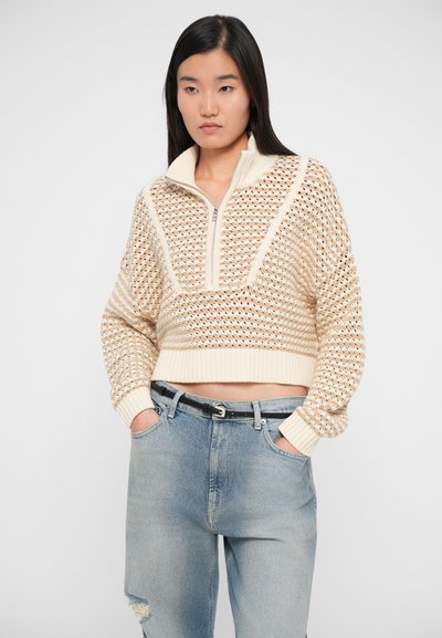 Knit pullover in cream and beige with a zippered collar, featuring an open weave pattern and ribbed cuffs. Worn with light blue distressed jeans.