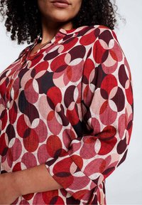 Donna che indossa una camicetta a maniche lunghe con un motivo circolare sovrapposto in rosso, rosa e bordeaux su uno sfondo chiaro e semplice.