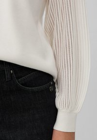 Wit langemouwen top met ribbelige doorschijnende mouwen, gecombineerd met donkerdenim jeans met een subtiel zakontwerp en metallic accenten.