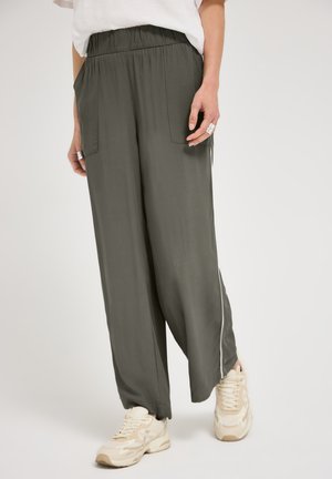 Femme portant un pantalon ample vert foncé avec une ceinture élastique, une bande blanche sur le côté, des poches avant, associé à des baskets beige et un haut blanc.