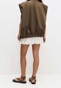 Brązowa kamizelka bez rękawów z elastycznym ściągaczem u dołu, zestawiona z białą falbanianą spódnicą i brązowymi płaskimi sandałami. Gładka tkanina, casualowy design.