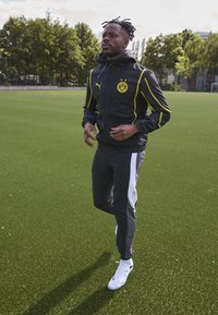 Chándal Puma en negro con detalles en amarillo, con un logo de BVB. La chaqueta tiene capucha; los pantalones son grises con una franja blanca. Zapatillas deportivas blancas.