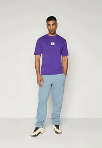 Purple t-shirt with a white logo on the chest, paired with light blue sweatpants featuring a black logo. The outfit is completed with black and white sneakers.