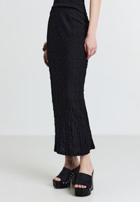 Jupe maxi noire plissée avec une finition texturée, associée à des sabots noirs épais avec des clous décoratifs et une semelle compensée.