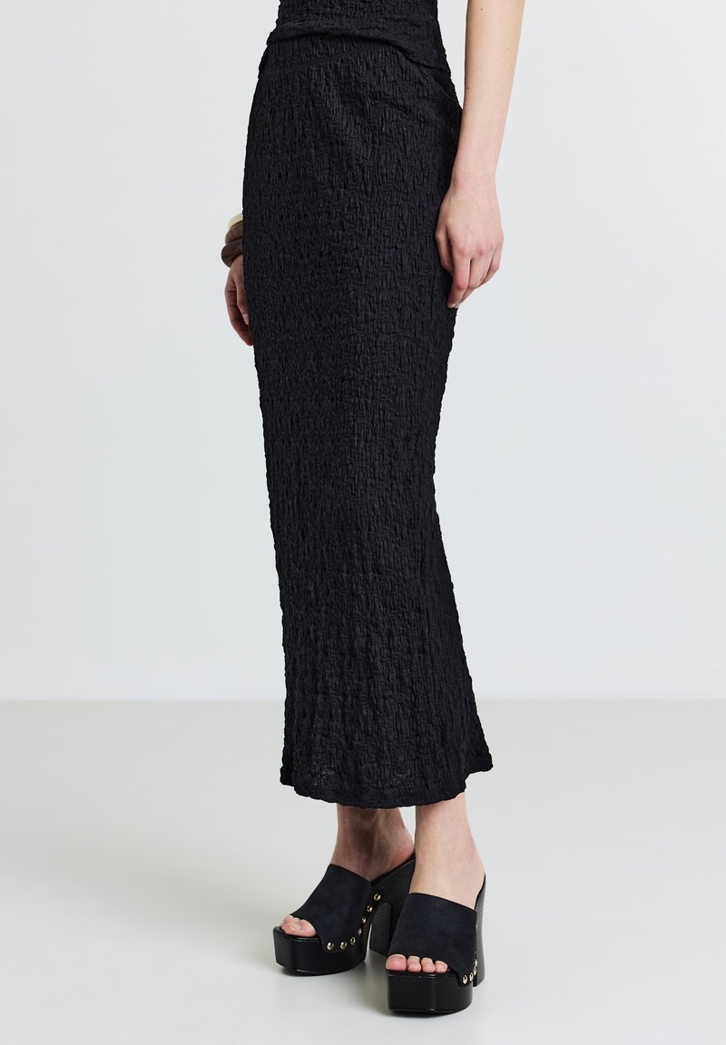 Schwarzer, zerknitterter Maxirock mit strukturiertem Finish, kombiniert mit klobigen, schwarzen Clogs mit dekorativen Nieten und einer Plattformsohle.