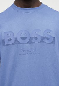 Κοντινό πλάνο ενός ατόμου που φοράει ανοιχτομπλε πουκάμισο με ανυψωμένα γράμματα "BOSS" και μικρότερο κείμενο "Tennis Club Established Sports Brand."