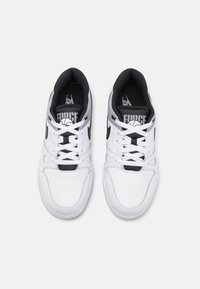 Baskets blanches avec des accents noirs, présentant une texture perforée, un bout rond et un système de laçage. Logo de la marque visible sur la languette.