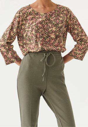 Kvinde iført en beige langærmet top med pink og grønt blomsterprint, parret med højtaljede olivengrønne bukser med snøre.