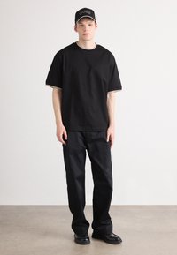 Camiseta negra de manga corta con costuras en contraste, combinada con pantalones negros y zapatos negros. Una gorra negra con un logo completa el conjunto.