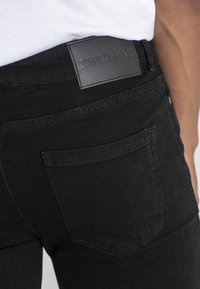 Musta farkkushortsit, joissa on sileä pinta, taustatasku ja musta nahkalappu, johon on kohokuvioitu "Gianni Kavanagh" vyötärönauhaan.