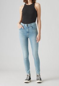 Jeans ajustados en denim azul claro, de cintura alta, con dos bolsillos delanteros, cierre de botón y costuras laterales. Combinados con una camiseta sin mangas negra y zapatillas.