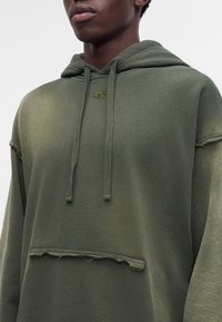 Jeune personne portant un sweat à capuche vert avec des coutures effilochées et un petit logo brodé sur la poitrine.