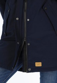 Granatowa kurtka z zamkiem z przodu i zatrzaskami, charakteryzująca się teksturowanym materiałem oraz skórzaną metką na dolnym brzegu.