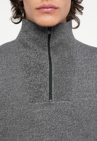 Pullover grigio scuro con colletto alto e chiusura a mezza zip. Tessuto a maglia con un motivo sottile e una zip nera in contrasto.
