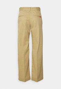 Pantalons en toile beige à jambes larges, offrant une texture lisse, deux poches arrière et un patch logo cousu sur la droite.
