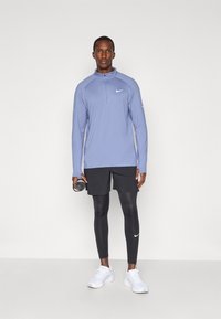 Lange mouwen, lichtblauw, rits sporttop met zwarte shorts en zwarte leggings. Witte sneakers en een fitnessaccessoire in de hand.