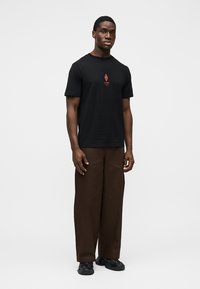 Homme debout portant un t-shirt noir avec un logo rouge, un pantalon marron à jambes larges et des baskets noires sur un fond blanc uni.