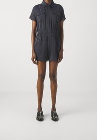 Osoba v černém krátkém overal s krátkým rukávem, předními kapsami a elastickým pasem, doplněná o černé loaferky s hroty, na jednoduchém pozadí.