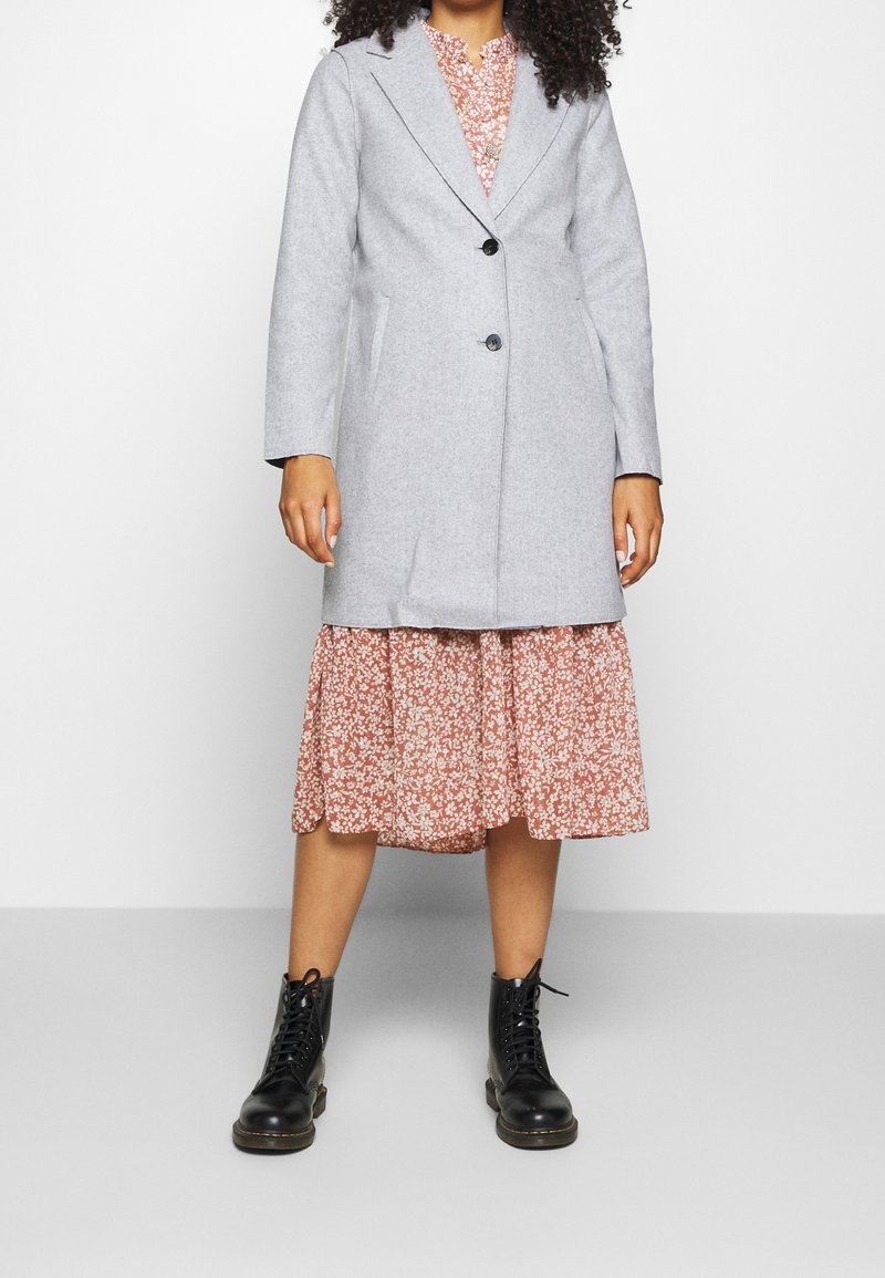 Vrouw in een lichtgrijze knoopjack over een bloemdress in roze met zwarte veterschoenen, staande tegen een witte achtergrond.