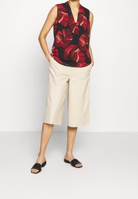 Kvinna som bär en ärmlös blus i svart och röd lövtryck, beige vida cropped byxor och svarta slip-on sandaler, står med händerna i fickorna.