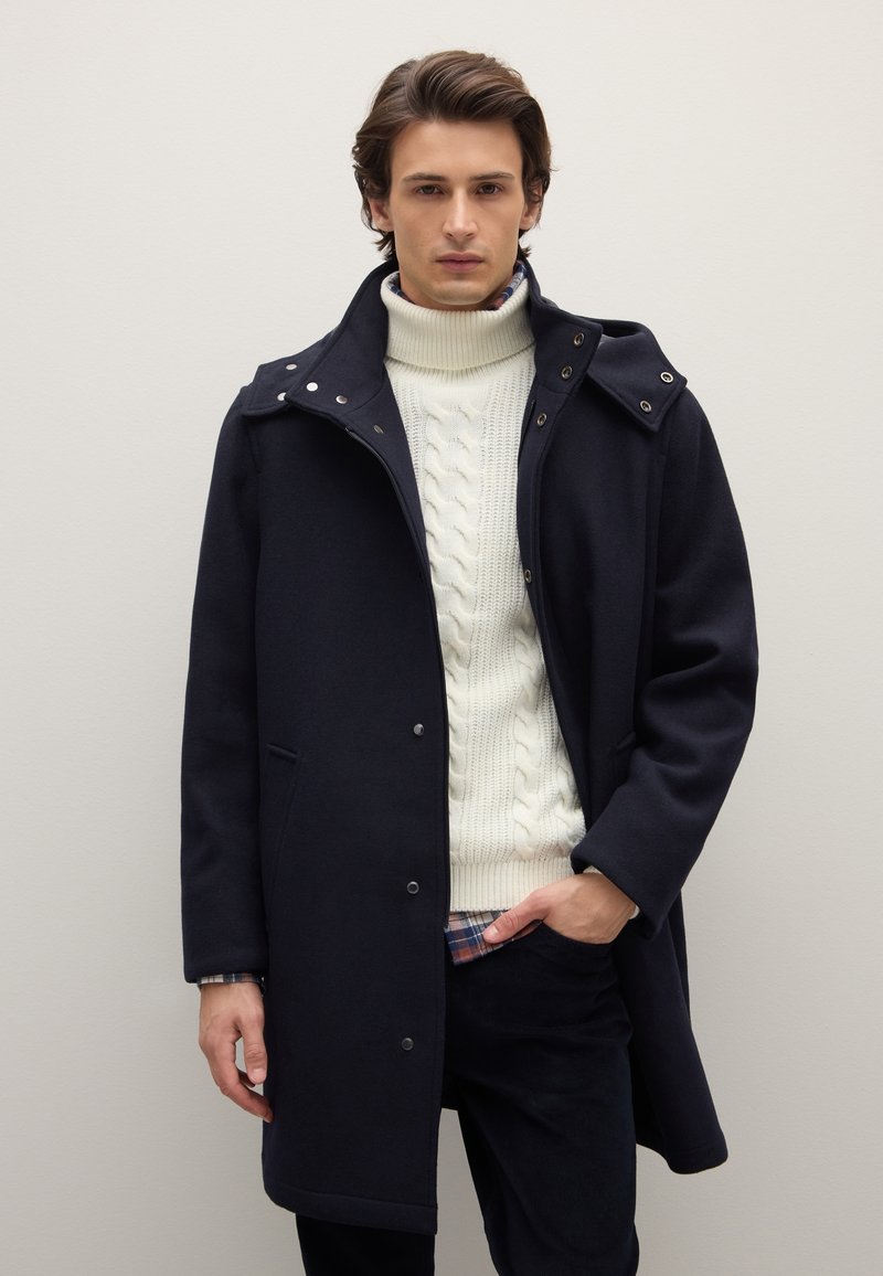 Cappotto blu navy in misto lana con colletto alto, chiusure a scatto e tasche laterali, indossato sopra un maglione crema a trecce.