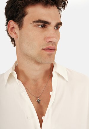 Collana con pendente d'argento a forma di croce, appesa a una sottile catena d'argento. Il modello indossa una camicia bianca a colletto con scollatura aperta.
