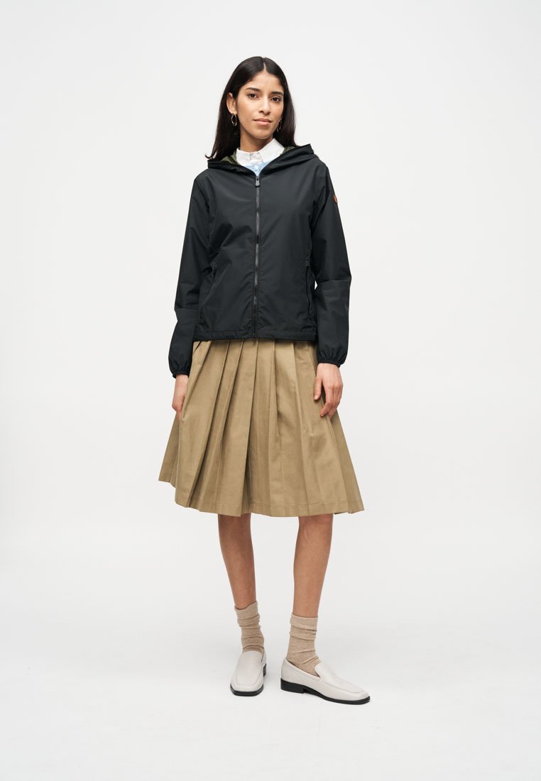 Femme debout portant une veste à capuche noire zippée, une chemise blanche, une jupe plissée beige, des chaussettes beiges et des mocassins blancs sur fond blanc.