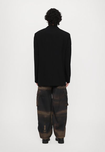 Schwarzes Blazer mit strukturierter Passform, kombiniert mit lockeren, braunen Cargo-Hosen mit einem Farbverlauf-Muster und Seitentaschen. Schwarze Schuhe runden das Outfit ab.