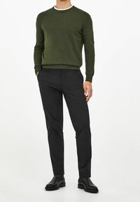 Olivgrüner Strickpullover mit weißem Rundhalsausschnitt, kombiniert mit schwarzen, schmal zulaufenden Hosen und schwarzen Leder-Stiefeletten. Einfaches und figurbetontes Design.