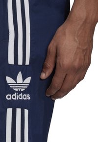 Námořnické modré sportovní kalhoty s bílým třípruhovým designem a vyšitým logem. Materiál vypadá hladce, ukazuje texturu a barevný kontrast.