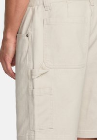 Beige cargo shorts med flere lommer, herunder en klaplomme på siden og en glat tekstur. Indeholder syede løkker til værktøj.