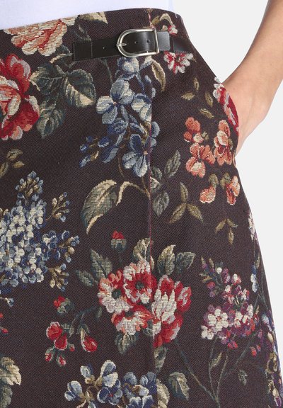 Jupe à motif floral avec des fleurs rouges, bleues et blanches sur un tissu foncé, avec une ceinture noire et une main dans la poche latérale.