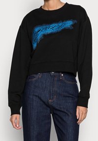 Persona que lleva un sudadera negra recortada con el logo de "Guess" en degradado azul y unos jeans de cintura alta de color azul oscuro con costuras naranjas.