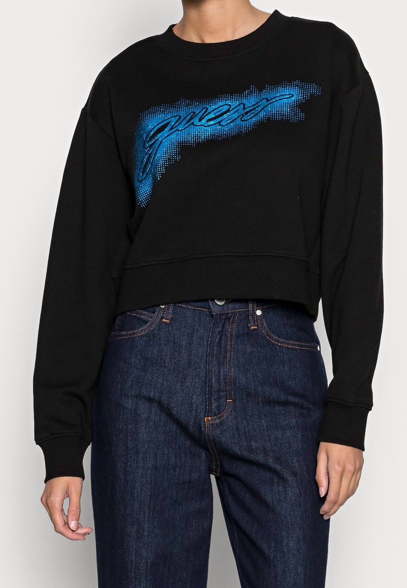 Persona que lleva un sudadera negra recortada con el logo de "Guess" en degradado azul y unos jeans de cintura alta de color azul oscuro con costuras naranjas.