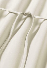 Helles beiges Gewebe mit glatter Textur, das ein geknotetes Bindungsdetail und leicht fallende Falten aufweist und aus einem weichen, elastischen Material besteht.