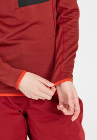 Rotes Langarmoberteil aus strukturiertem Stoff, mit kontrastierenden orangenen Akzenten an den Bündchen und einem schwarzen Patch auf der Brust, kombiniert mit roten Hosen.