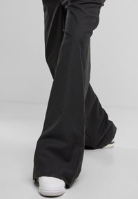 Personne portant un pantalon noir ample évasé et des baskets blanches, les jambes croisées, debout sur un sol gris clair avec un fond uni.